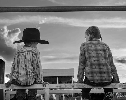 A photograph of two children sitting on a fence wearing western wear, looking away from the camera, nearly silhouetted against the sky.