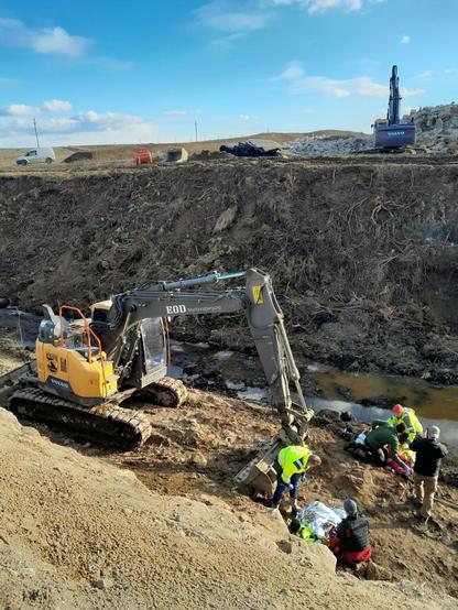 The picture shows an exercise in which an injured person is being rescued. Two excavators can be seen in the background, one of which is labelled ‘EOD ammunition recovery’. The exercise simulates a rescue in connection with construction work or ground work. Several people gathered around an injured person wrapped in a silver rescue blanket.