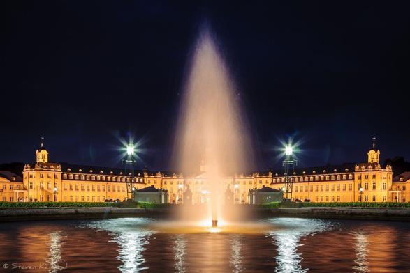In the foreground is the pool of water with a fountain spraying above it. It is a night scene and there are lights reflected in the pool and illuminating the fountain water droplets. There is a large castle in the middle ground, with a turret on either end. It is fully lit and the sky is black above it