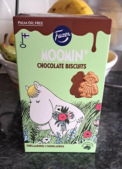 A green box containing biscuits shaped like characters from the Moomintroll books. The biscuits are half coated in chocolate. The front of the box has an illustration of Snorkmaiden, who is picking flowers.