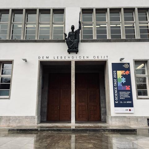 Entrance of the 'Neue Universität' building of the University of Heidelberg: A wooden two-door entrance with a statue above (with the letters "DEM LEBENDIGEN GEIST" underneath) and a banner of the conference E-Science-Tage 2025.