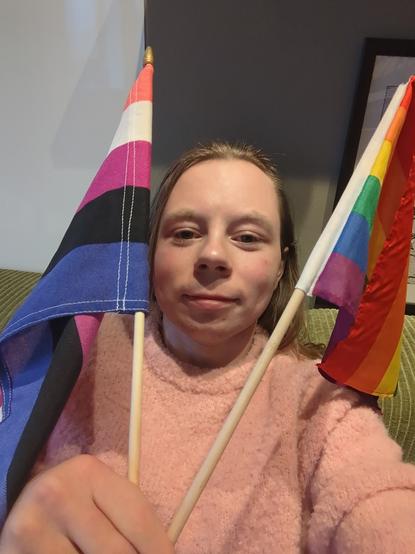 A woman is seen holding the pride and gender-fluid flag.

She is sitting on a couch, wearing a pink sweater!