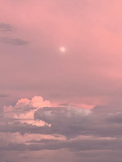 A tiny glowing moon softened by cloud cover in a hazy pink sky with gray and pink clouds below.