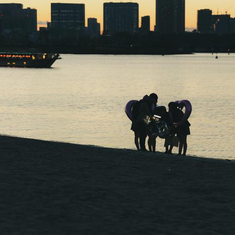 The image depicts a group of people standing near the water's edge at dusk, with a city skyline in the background. They appear to be holding or surrounded by inflatable rings or balloons, possibly shaped like hearts. The lighting is low, suggesting a sunset scene.