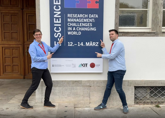 Tabea Tietz (left) and Jonatan Jalle Steller standing left and right of the advertisement poster for the E-Science Tage 2025 in Heidelberg pointing at tge poster with their fingers. The poster is located left of the entrance of the Heidelberger Universität. Both are waering a light blue colored shirt and dark pants.