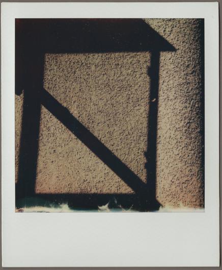 Polaroid of the shadow of an outdoor wall lamp makes a square with a diagonal through it against a brown stucco wall.