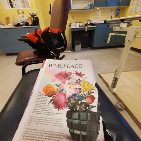 The photograph shows a book titled "WAR/PEACE" resting on the dark, padded seat of a chair. The book's cover features an image of a vase shaped like a hand grenade, overflowing with colorful flowers. The words on the book discuss topics related to social issues and conflict. The chair is situated in what appears to be a medical or therapy office, suggested by the presence of medical equipment, cabinets, and anatomical posters in the background. Miscellaneous objects and equipment occupy the counters around the chair.