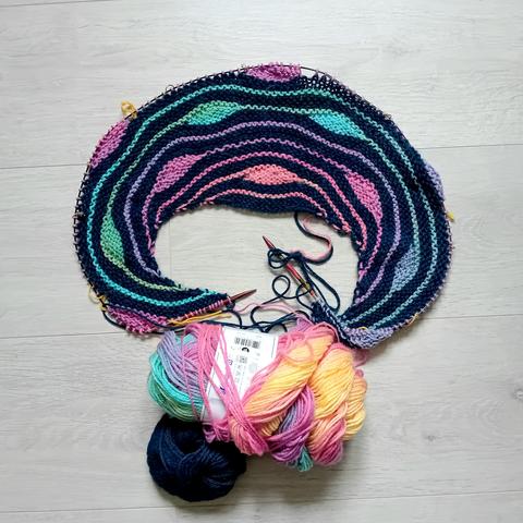 Chale arrondis en cours de tricot. Il est rayé arc-en-ciel sur fond bleu canard. Sur les lignes arc-en-ciel, il y a des gouttes horizontal à intervalle régulier.