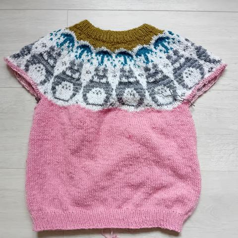 Pull tricoté en cours. L'encolure présente une frise avec le personnage totoro répéter plusieurs fois. La couleur principale est rose, mais le col est vert olive. Il reste les bras à tricoter