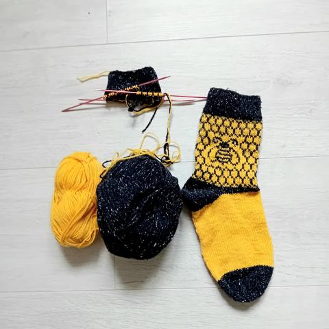 Chaussettes jaunes et noirs tricotées. La première est finie. Il y a une abeille en jacquard sur des alvéoles de ruches. Le talon  et la pointe sont noire. La deuxième chaussette est à peine commencé. Le dessin n'est pas encore visible.