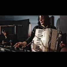 A long-haired, clean-shaven musician wearing glasses and a long-sleeved shirt is on stage, playing a synthesizer keyboard while wearing a piano accordion.