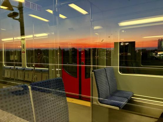 Das Bild zeigt das Innere eines Zuges bei Sonnenuntergang. Im Vordergrund sind blaue gepolsterte Sitze zu sehen. Im Hintergrund, durch die Fenster des Zuges, ist ein lebendiger Sonnenuntergang zu sehen, der den Himmel in Orange- und Rottönen malt. Die Lichter der Stadt sind in der Ferne sichtbar. Die Reflexionen im Fenster vermischen das Innere des Zuges mit der Außenansicht. Eine rote Tür im Zug ist ebenfalls sichtbar. Die Gesamtatmosphäre ist ruhig und friedlich.

Bereitgestellt von @altbot, generiert mit Gemini