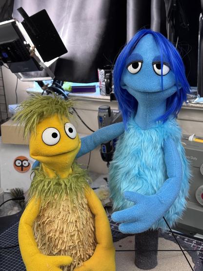 Two pointy-headed puppets, one with yellow fur and short green hair, and a blue one with sleepy eyes and long blue hair