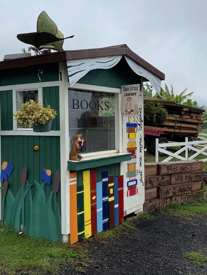 Cute Little Free Library on the premises of a small farm in Maui. The shed that houses the library is decorated with books and wildflowers, and there is a small statue of a mongoose reading a book in the front.