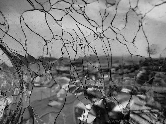 Una imagen en blanco y negro muestra una superficie de cristal de un jarrón, con efecto de agrietado. Las grietas se parecen a una red compleja  creando un patrón orgánico. A través de las grietas se puede ver la ciudad como un fondo borroso. El enfoque principal está en el patrón de las grietas del cristal, creando una textura fragmentada.