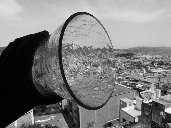 Una mano sostiene una pieza de vidrio transparente con forma de cuenco invertido. El vidrio está agrietado en una red de finas líneas, creando un efecto artístico. A través del vidrio, se ve un panorama de una ciudad, con edificios de varios pisos y un horizonte montañoso al fondo. La imagen está en blanco y negro, lo que le da un aspecto dramático y monocromático. La mano y el vidrio son el foco principal, con la ciudad como telón de fondo.