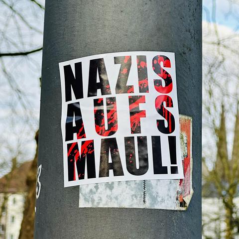 Auf einem Aufkleber an einem Laternenpfahl steht in fetter schwarzer Schrift "NAZIS AUFS MAUL!" vor einem weißen Hintergrund. Die schwarze Schrift hat rote Spritzer, was die Antifa-Botschaft optisch unterstreicht