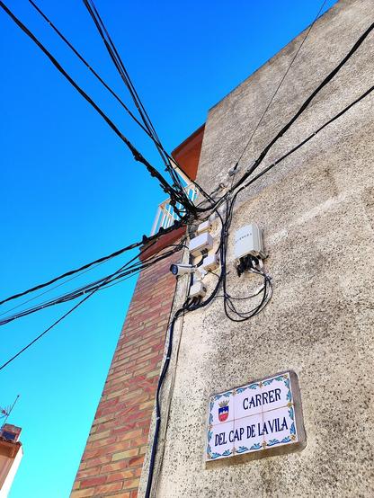 Rètol de ceràmica decorada amb el nom 'Carrer del Cap de la Vila' i l'escut de la Serratella en la façana d'una casa. A sobre, una càmera de seguretat, caixes de connexió elèctrica i cables. Al fons, el cel blau i ras.