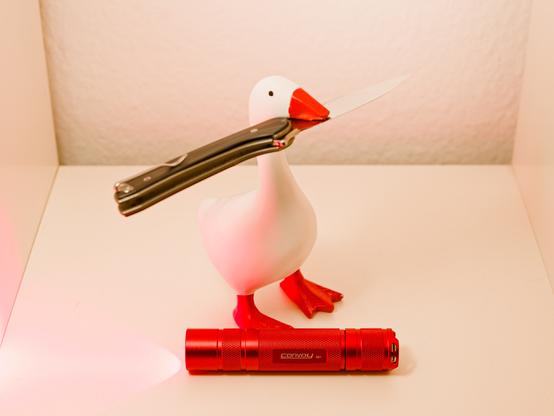 A red colored Convoy S2+ flashlight emitting red light sits below a goose figurine holding an opened pocket knife