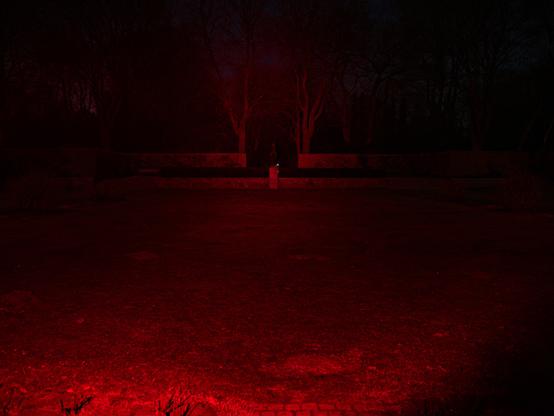 A field in a park with a horse and rider statue at the far end with walls and trees beyond, lit in red
