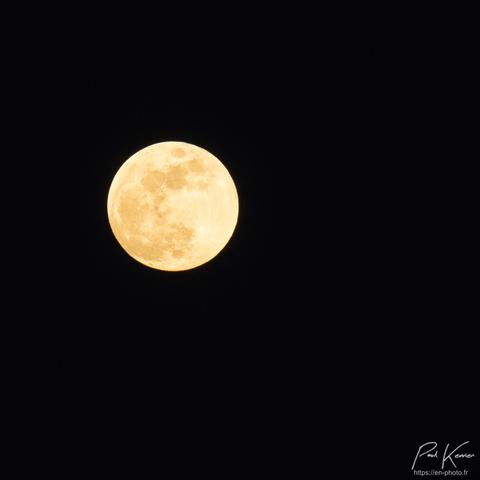 Photo en couleur de format carré, présentant une pratiquement pleine lune dans un ciel sombre avec quelques détails visibles, mers, cratères...