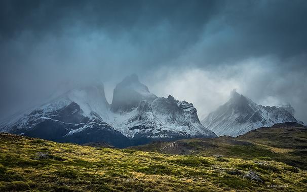 Cloud filled skies obscure chiselled, snow dusted mountain peaks.  Sunlight illuminates the grassy foreground providing a juxtaposition in the landscape.