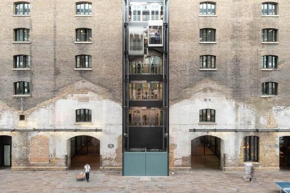 19th century brick industrial building converted to a library. The view is taken across an interior passage, the facade in the backdrop shows traces of former industrial sheds. A bank of modern elevators are in motion against it.