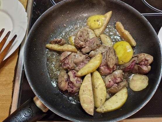 Na zdjęciu widać patelnię z przygotowywanym daniem. W skład potrawy wchodzą kawałki wątróbki oraz plastry jabłek, które są smażone razem na tłuszczu. Danie znajduje się na kuchence elektrycznej, a w tle widoczne są drewniane sztućce oraz talerz.