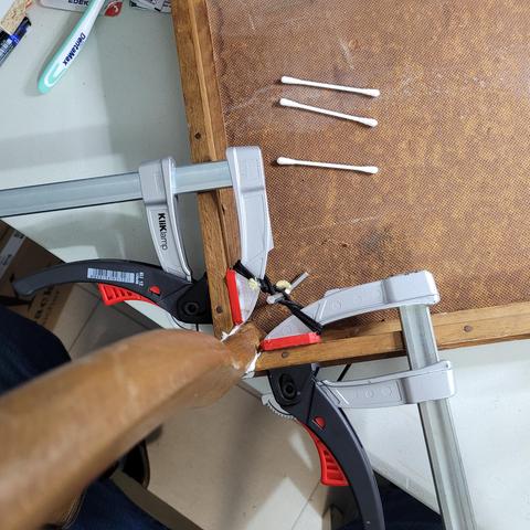 Das Bild zeigt eine handwerkliche oder restaurative Arbeit an einem Holzrahmen, der mit mehreren Schraubzwingen fixiert wurde. Die Schraubzwingen (Marke „KLIKlamp“) halten den Rahmen stabil auf einer Tischplatte.

Besonders auffällig ist die schwarze Schnur mit gelbem Kern, die mit einem Knoten um einen Metallstab gewickelt wurde. Diese Konstruktion könnte eine Spann- oder Haltefunktion haben. Zusätzlich sind drei Wattestäbchen auf der Oberfläche zu sehen, was darauf hindeuten könnte, dass Klebstoff oder eine andere Substanz aufgetragen oder gereinigt wurde.

Ein Holzstab oder Werkzeug mit einer weißen Substanz an der Spitze (vermutlich Leim) ragt ins Bild, möglicherweise um eine Reparatur an der beschädigten oder gelösten Holzverbindung vorzunehmen. Das gesamte Setup wirkt wie eine improvisierte Spannvorrichtung für eine präzise Reparatur oder Restaurierung des Rahmens.