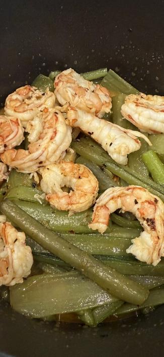 En la imagen se observa un plato de camarones a la plancha acompañados de borrajas cocidas. Los camarones tienen un aspecto jugoso y ligeramente dorado, con un toque de especias que resalta su color y textura. Las borrajas, de un verde intenso y brillante, parecen haber sido cocidas hasta alcanzar una textura tierna, posiblemente con un poco de aceite y condimentos. El conjunto está servido en un recipiente oscuro, probablemente una sartén o un wok, lo que resalta los colores y la frescura de los ingredientes.