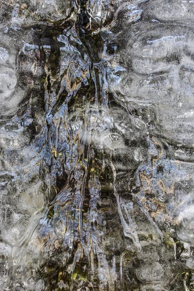 A layer of ice that  reveals the rock and moss patterns behind the ice and the patterns within, while also reflecting the blue sky and dark tree branches above it. These patterns and colors combine and intersect to form imagery befitting the imagination.