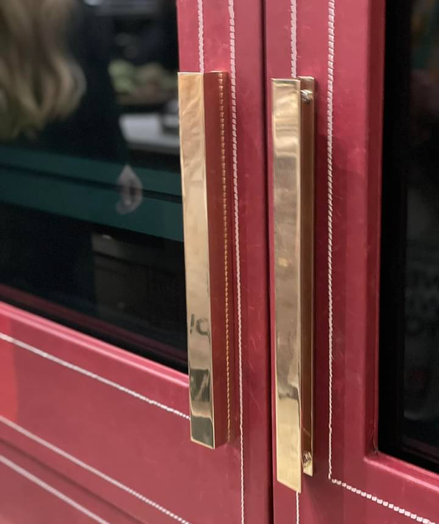 Close-up view of two brass handles on a glossy red double-door appliance, possibly a refrigerator or oven. The red doors have decorative white stitching-like lines, giving an elegant touch. The blurred background shows a person with long hair and some kitchen items, suggesting a kitchen environment.