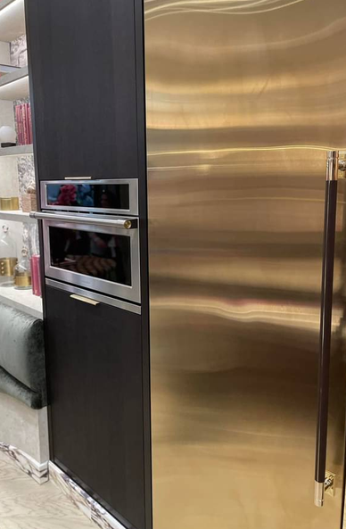 Modern kitchen area featuring a dark-toned cabinet with a built-in stainless steel oven. To the right, a large, gold-colored refrigerator with a vertical handle stands prominently, wrapped in a dark leather. Above the cabinet, open shelves display decorative items like books, a white globe, and a gold vase, adding a stylish and personal touch. The kitchen floor showcases a marble-like pattern, completing the sleek and elegant aesthetic.