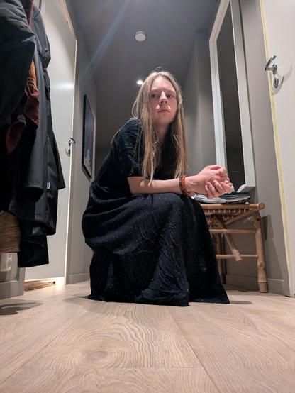 The image captures a woman in her apartment. She is sitting on the floor.

In the background, coats hang on a rack, a wooden bench, and a painting adorns the wall, and the stark lines of doorways create a sense of depth.