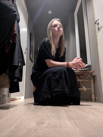 The image captures a woman in her apartment. She is sitting on the floor.

In the background, coats hang on a rack, a wooden bench, and a painting adorns the wall, and the stark lines of doorways create a sense of depth.