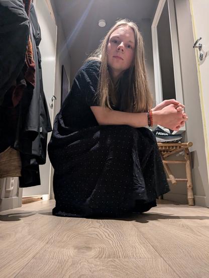 The image captures a woman in her apartment. She is sitting on the floor.

In the background, coats hang on a rack, a wooden bench, and a painting adorns the wall, and the stark lines of doorways create a sense of depth.