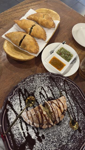 This photo showcases a meal featuring both savory and sweet empanadas.  

In the upper part of the image, there is a wooden plate holding three golden-brown, deep-fried savory empanadas resting on a napkin. They appear crispy and have been cut open, revealing a filling that seems to include vegetables or meat. Accompanying them on a white plate are two dipping sauces—one green and chunky, possibly a salsa verde, and another orange-red, likely a spicy sauce.  

In the lower portion of the image, a dessert empanada is plated on a dark dish with an artistic drizzle of chocolate sauce and a dusting of powdered sugar. The empanada has been cut in half, revealing a warm, possibly fruit-based filling. Two golden spoons accompany the dessert, adding an elegant touch.  

The food presentation is rustic yet refined, served on wooden and ceramic plates, and is placed on a wooden table. The contrast between the savory and sweet empanadas makes for an appealing meal.