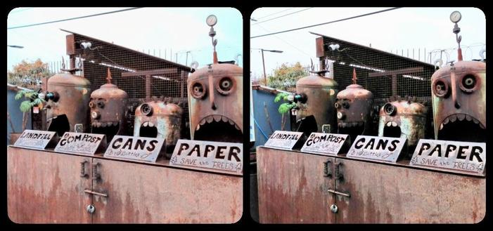 Four rusty propane tanks turned into faux robot heads over a recycling bin. They are labeled Landfill, Compost, Cans, and Paper.