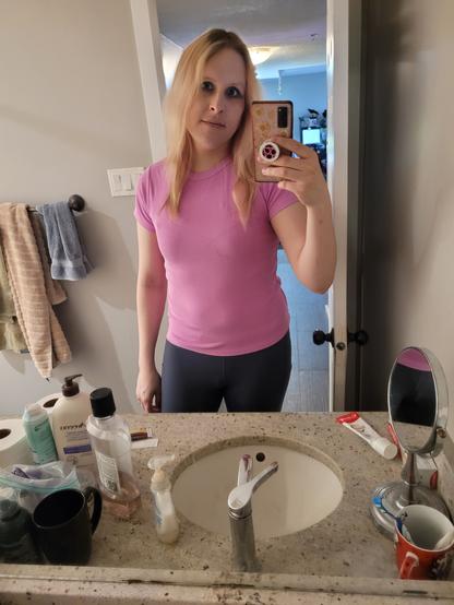 trans girl bathroom mirror selfie. she's wearing a pink shirt and blue leggings. her blue eyeshadow darkens her eyes a little bit. her hair is bleach blonde with aged out pink dye and brown roots showing