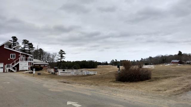 Acres of fields under a cloudy sky. The fields are pale gold. There are some red outbuildings and a white pen that's empty.