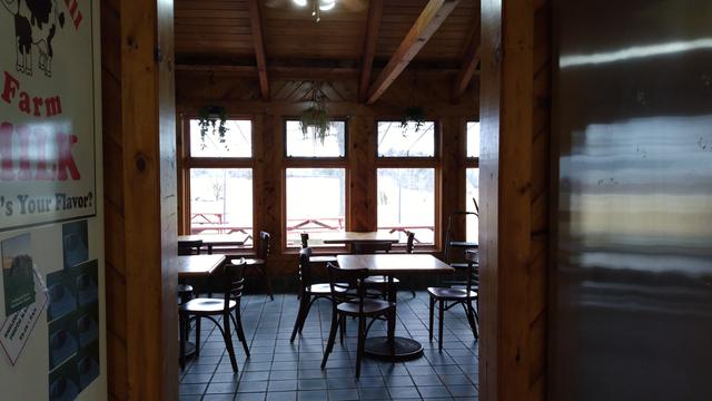 The inside of an ice-cream parlor, overlooking the fields. The parlor is empty, but soon will be filled with folks eating ice cream. The interior is wooden, and the table and chairs are cafe-style.