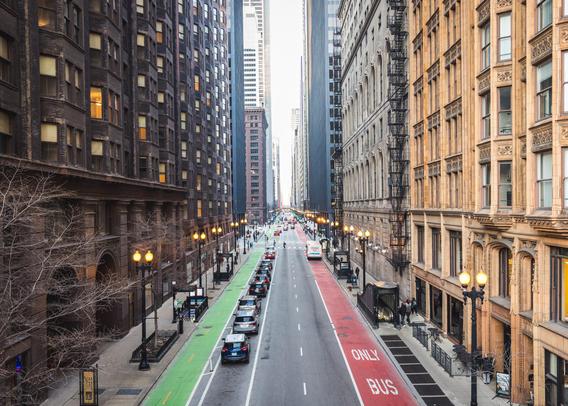 A city street is lined with tall buildings, showcasing a mix of architectural styles. The road has designated bike lanes and a bus-only lane marked in red. Several parked cars are on the street, and a few pedestrians are walking along the sidewalk.