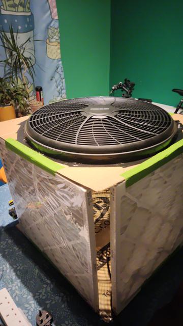 A partially constructed Corsi-Rosenthal Box. This is a cube, around 20 inches per side. The bottom is just cardboard. The top is a round black plastic fan, mounted in a hole in the middle of another piece of cardboard, and held there with black duct tape. The four sides are filter boards - cardboard frames containing cloth like filter material. The boards are attached to the top and bottom with green masking tape. There are gaps at the corners because the boards are smaller than the fan's diameter.