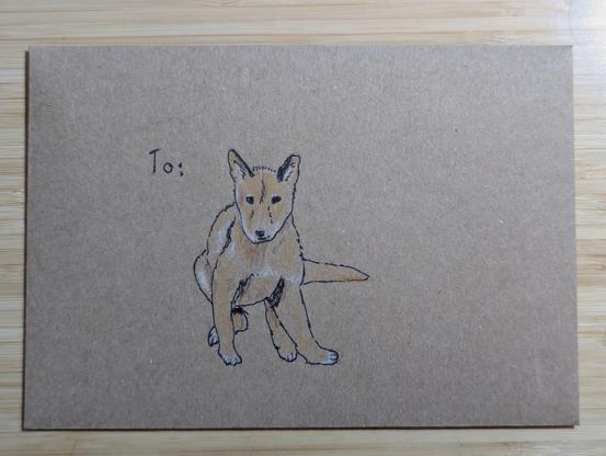 A recycled paper envelope with the word "To", followed by a pen & coloured pencil sketch of a dingo puppy sitting down with its legs sprawled under it