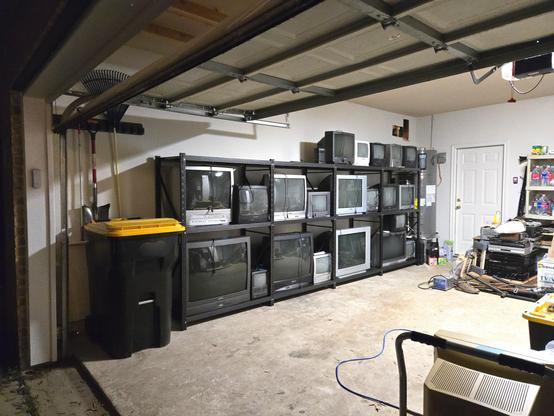 Photo of a set of metal shelves filled with different sizes of CRT televisions as viewed from just outside a raised garage door.
The shelving rack takes up most of a wall inside this garage, with only enough remaining space for a trash can at one end and a water heater tank at the other.
More to the center of the garage, a CRT computer monitor sits on a dolly and a wooden pallet supports three stacks of media players - DVD, VHS, 8 Track, etc.