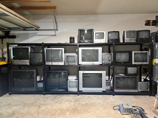 Photo of the same set of metal shelves filled with various sizes of CRT televisions, but this time, they are viewed straight-on from the opposite wall of the garage.
There are 22 TV sets varying in screen size from 9 inches to 36 inches, and some of them have built-in DVD players or VCR units.