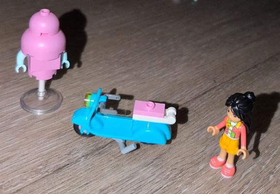 The image contains three LEGO toys: a pink and white robot on a stand, a blue scooter, and a girl with a pink shirt and orange shorts. The scooter is in the middle of the image, and the robot and the girl are on either side of it. The image is taken from a slight angle, and the background is a wooden surface. The text below the image says "The scooter is a compact LEGO model with a bright bl... 230.79 KB image." This suggests that the image is part of a larger collection of images depicting LEGO toys.