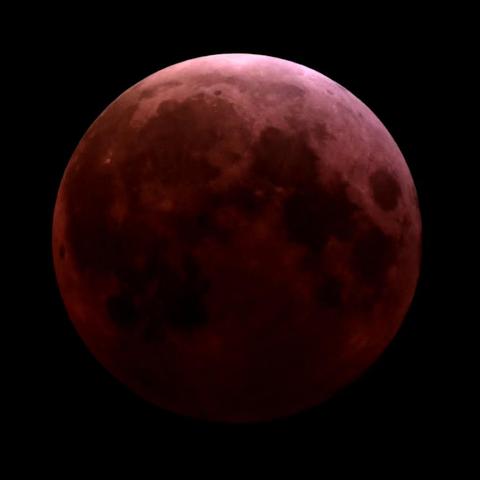 A close-up telescope shot of the fully eclipsed Moon. It appears be a deep red. Details of the moon's surface including the craters and dark maria are somewhat clear through the red cast.