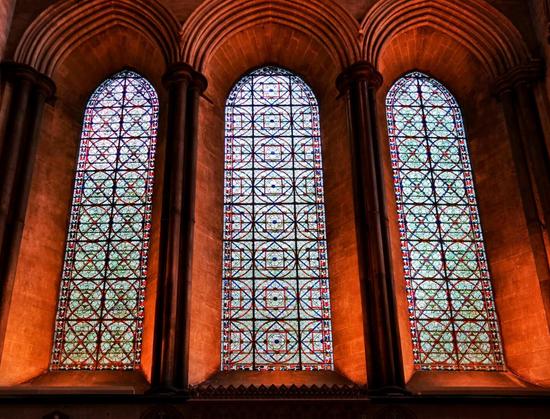 Three tall, arched stained glass windows are set in a dark stone wall. The windows are filled with a geometric pattern of small, colourful glass pieces, predominantly blue, green, and red. The stone arches above the windows are deeply recessed and the lighting is dim, casting shadows on the stone and highlighting the intricate patterns of the stained glass.
