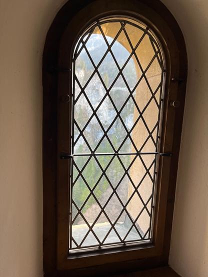 Das Fenster ist schmal, hoch und oben rund. Es hat einen braunen Holzrahmen und hat außen ein schwarzes X-Gitter. Draußen erkennt man Bäume und etwas braunen Weg und Berge.